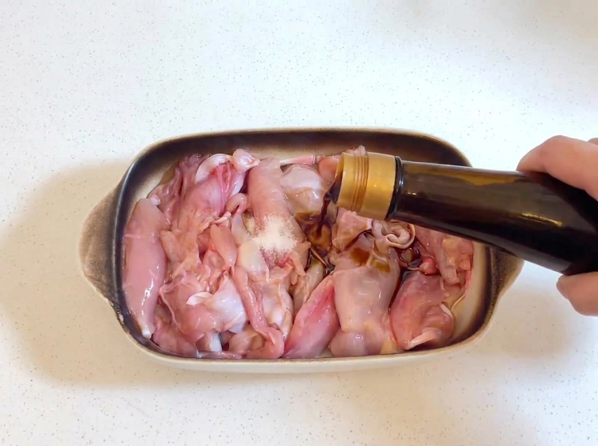 Salsa de soja oscura siendo vertida desde una botella de vidrio sobre trozos de pollo crudo, con una pequeña pizca de sal visible en el pollo.