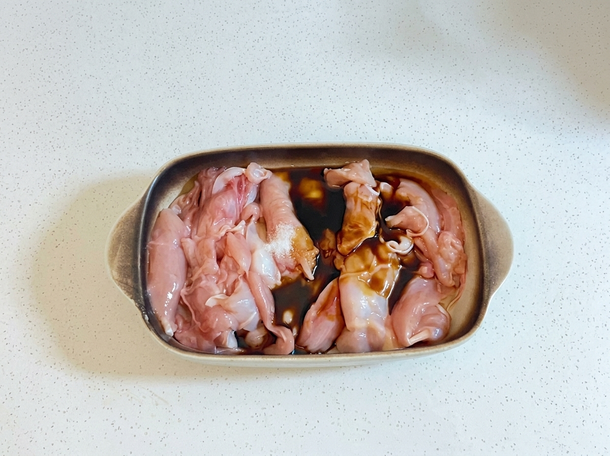 Líquidos de cocina oscuros y aceite siendo vertidos sobre trozos de pollo crudo en una fuente para hornear.