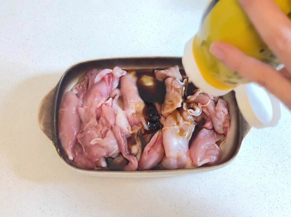 Salsa de ostras oscura y espesa siendo exprimida desde una botella amarilla sobre trozos de pollo crudo que reposan en un marinado de salsa de soja.