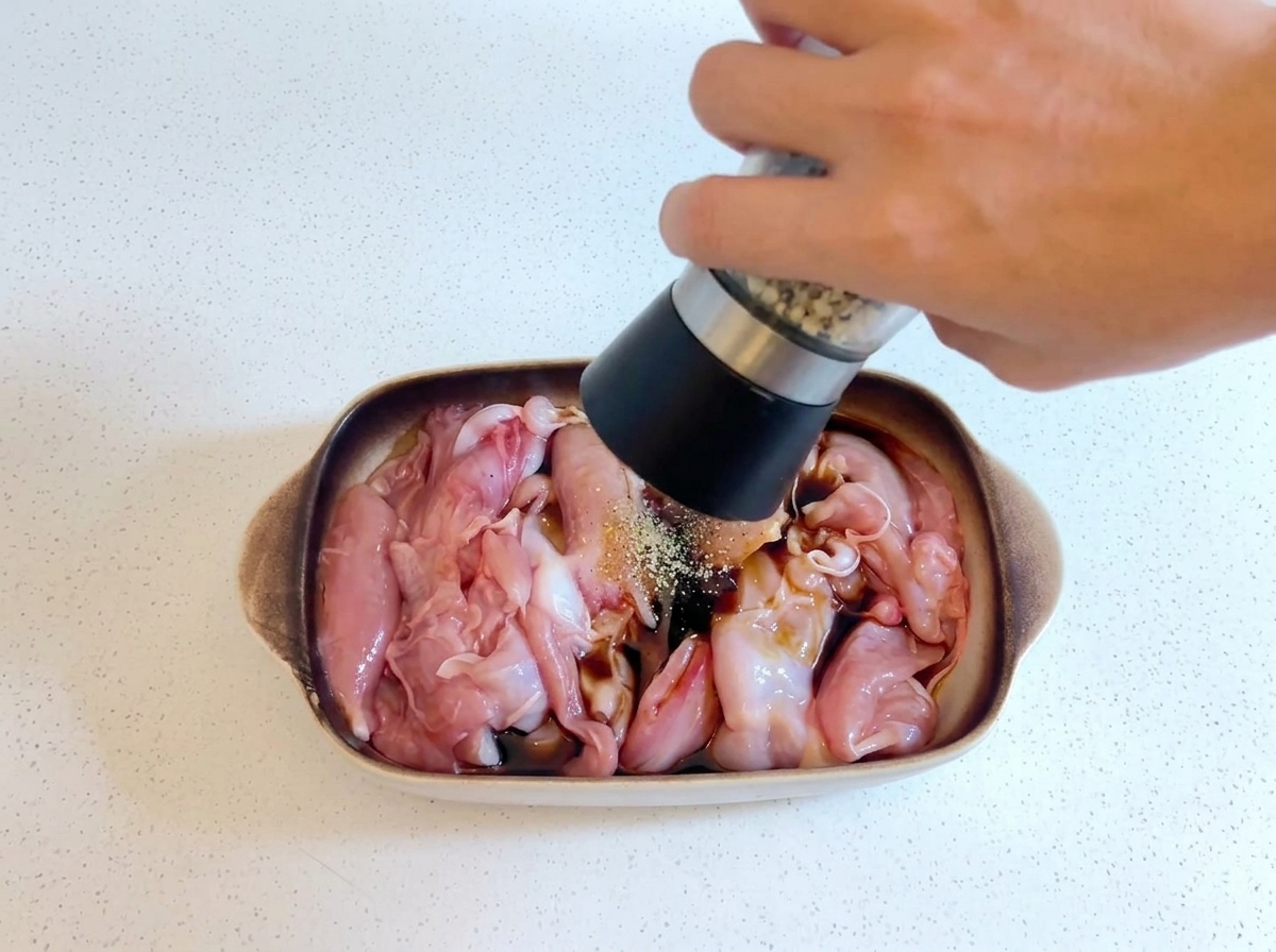 Una mano sosteniendo un molinillo de pimienta y moliendo pimienta fresca sobre el pollo crudo que se marina en un plato.