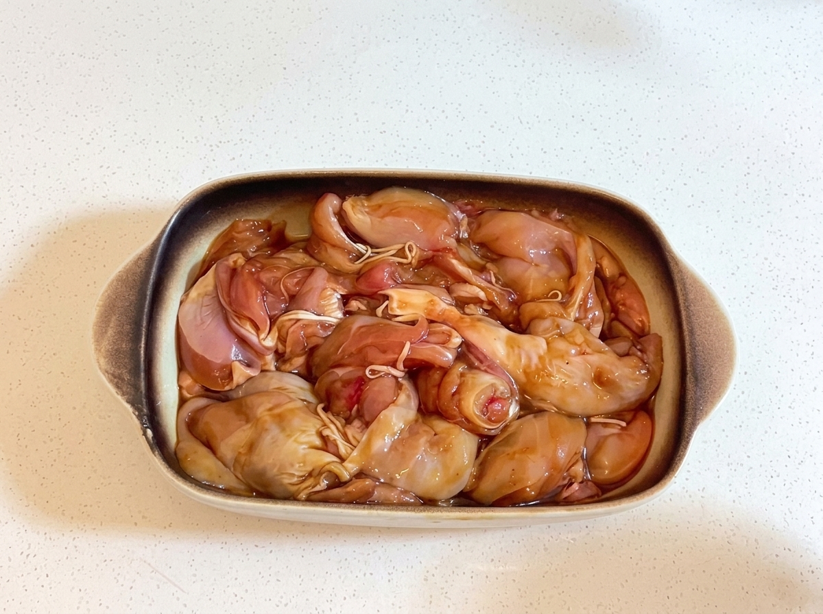 Una fuente de cerámica rectangular para horno llena de trozos de pollo marinados, firmemente cubierta con film plástico transparente.