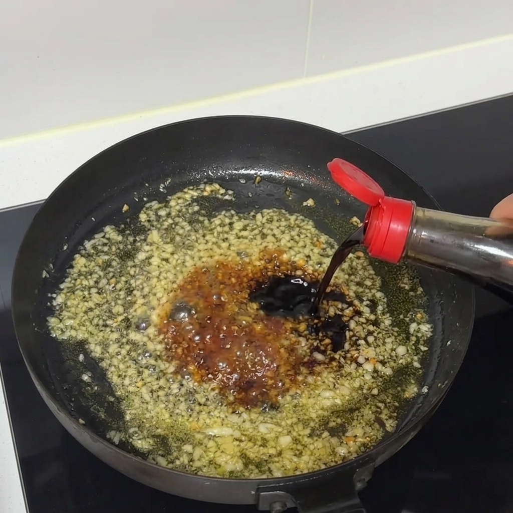 Salsa de soja oscura cayendo desde una botella hacia una sartén chispeante de ajo picado y salsa.