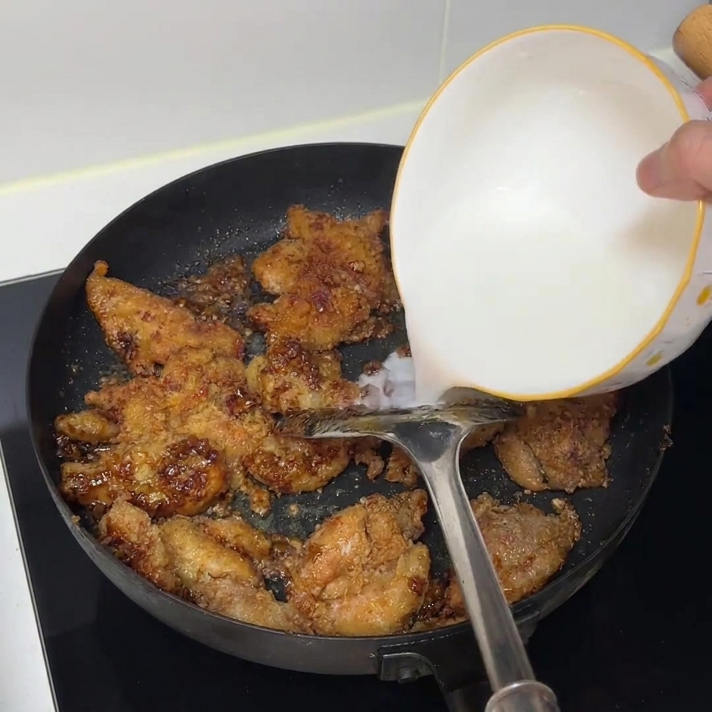 Agua con almidón siendo vertida desde un bol blanco en una sartén llena de trozos de pollo cocinado.