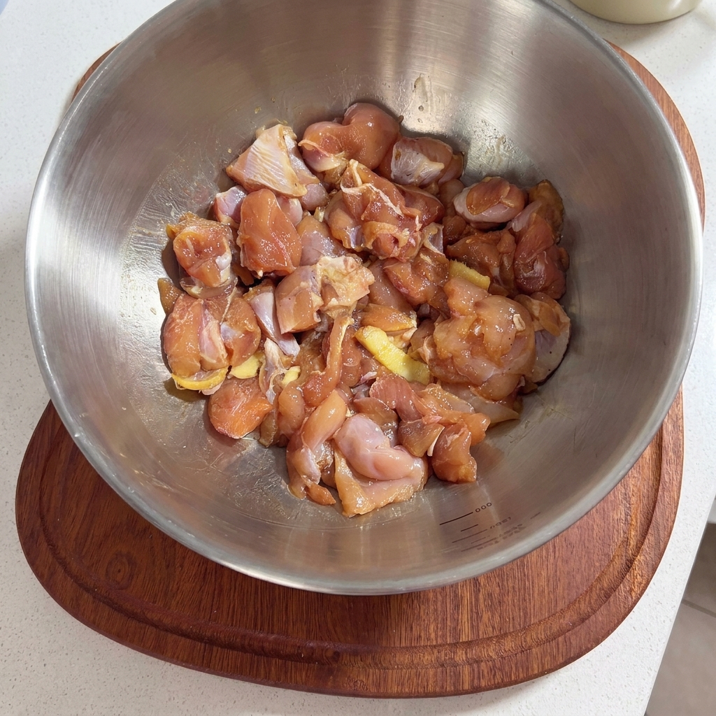 Un bol de metal lleno de trozos de muslo de pollo crudos sazonados con salsa de marinada y rodajas de jengibre.