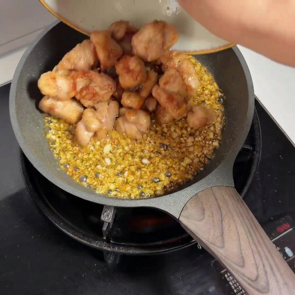 Trozos de pollo prefritos siendo vertidos desde un plato a una sartén llena de salsa de ajo.