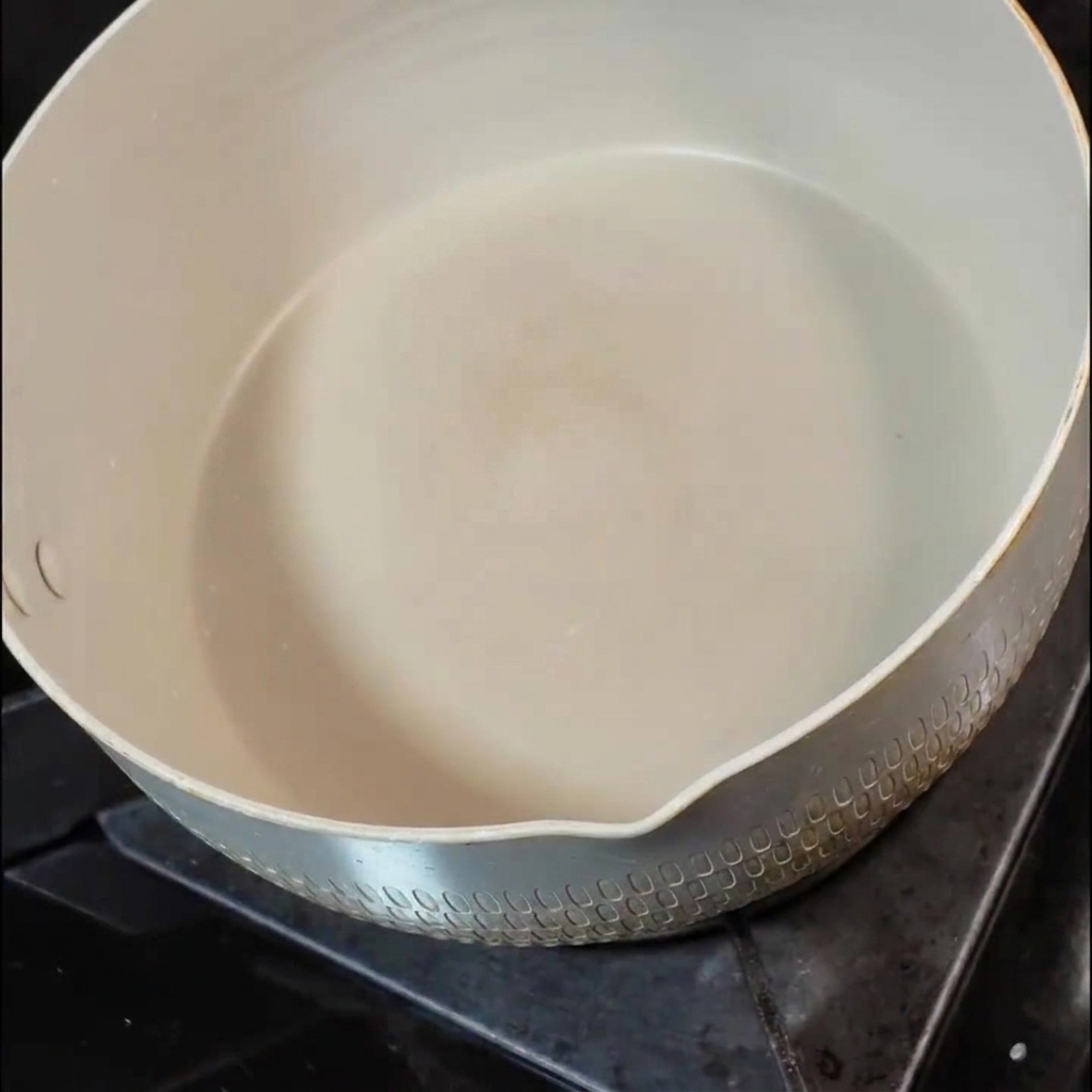 Un pequeño trozo de mantequilla derritiéndose dentro de una sartén de cerámica blanca en una hornalla de la estufa.
