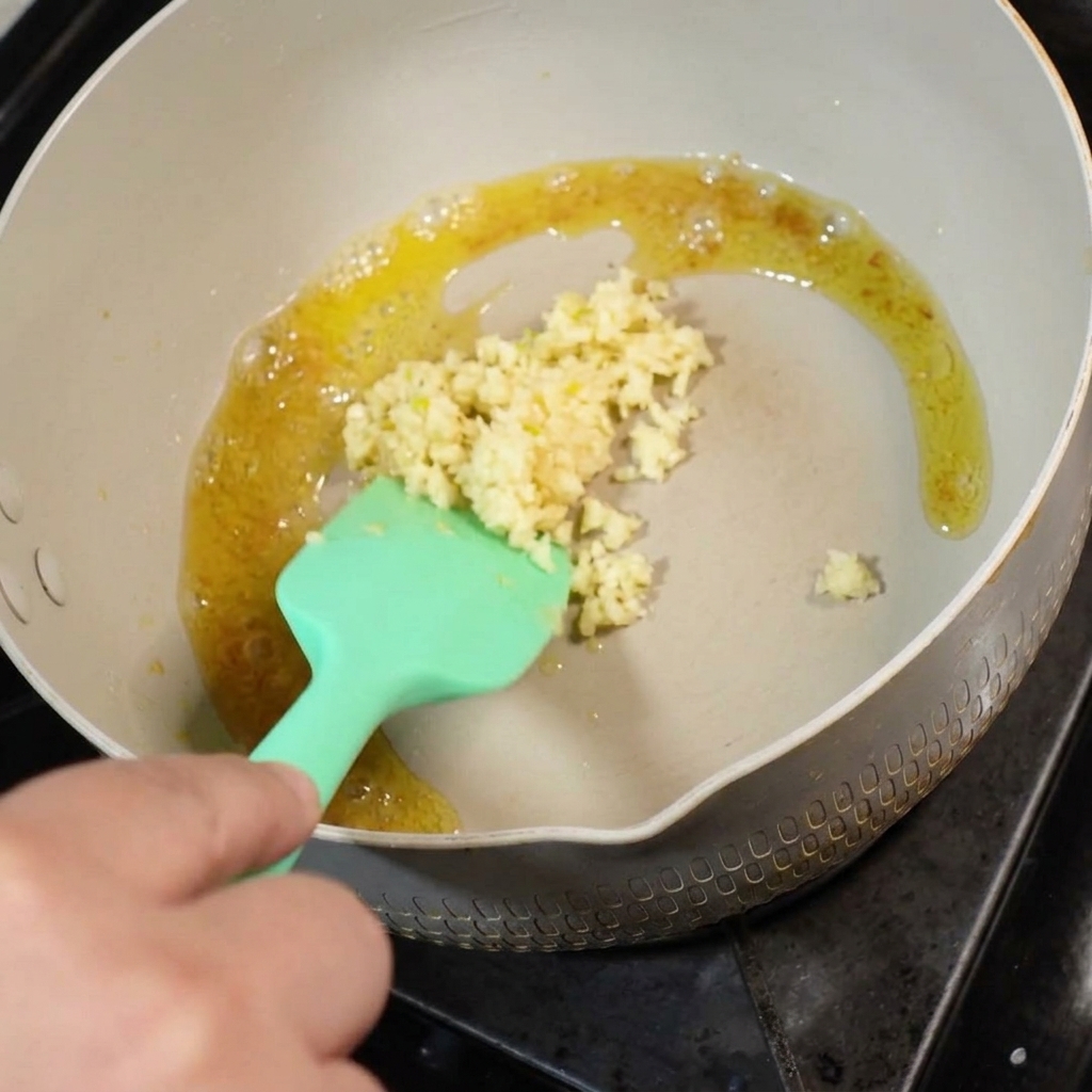 Ajo picado fresco siendo añadido a la mantequilla derretida en una sartén, revuelto con una espátula de silicona verde.