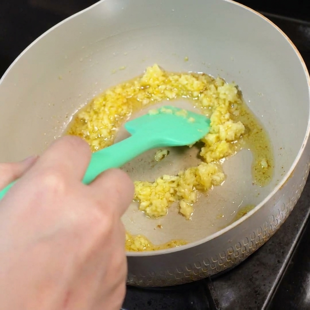 Ajo picado friéndose en mantequilla derretida en una sartén, revuelto con una espátula de silicona verde.