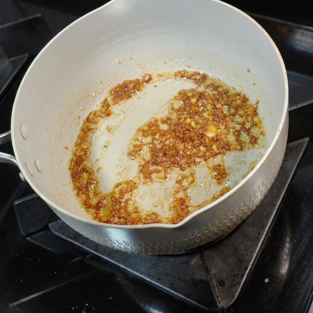 Un cucharón pequeño de caldo de fideos sabroso siendo vertido en la mezcla aromática de mantequilla y ajo.