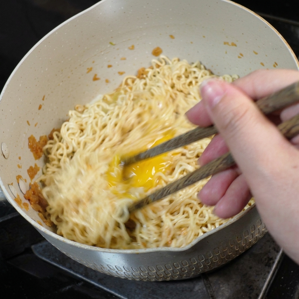 Un huevo siendo revuelto e integrado en los fideos dentro de una sartén usando palillos de madera.