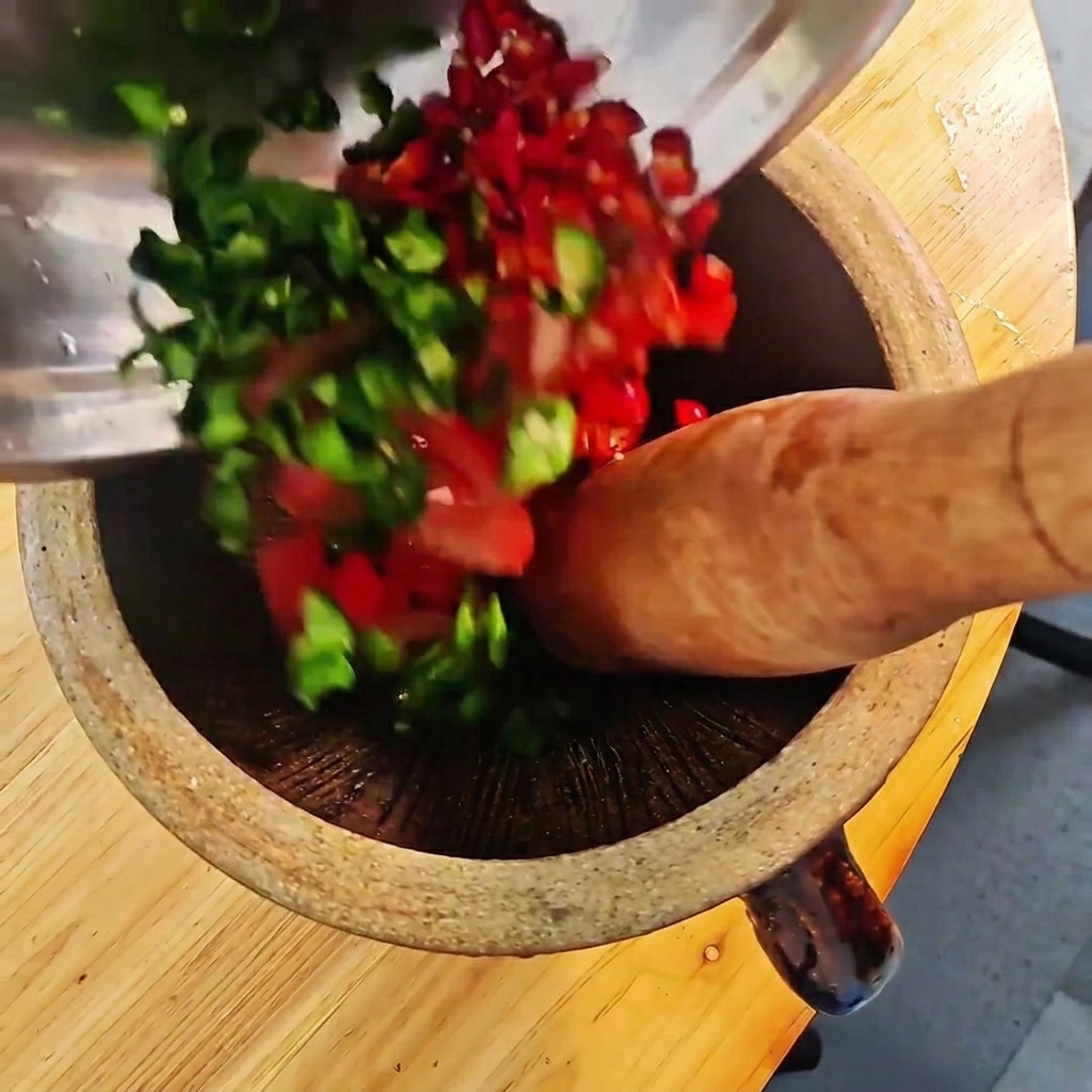 Chiles verdes y rojos picados y tomates siendo triturados en un mortero de madera con un mazo.