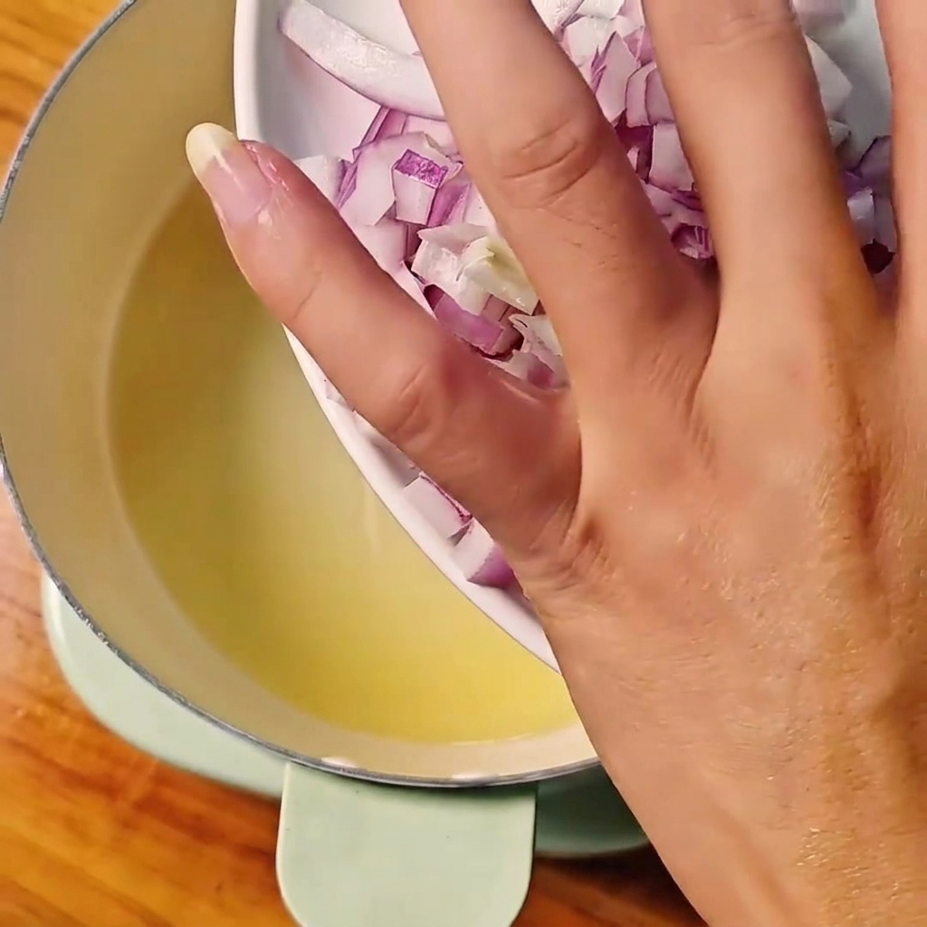 Cebollas rojas en dados siendo vertidas desde un plato blanco en una olla con aceite caliente.