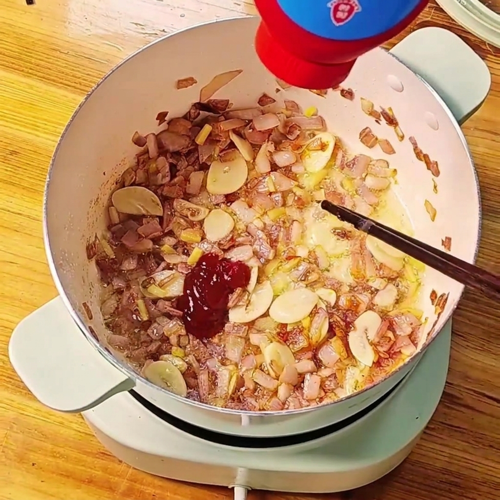 Pasta de tomate roja siendo exprimida de un tubo hacia una olla con cebollas y ajo sofritos.