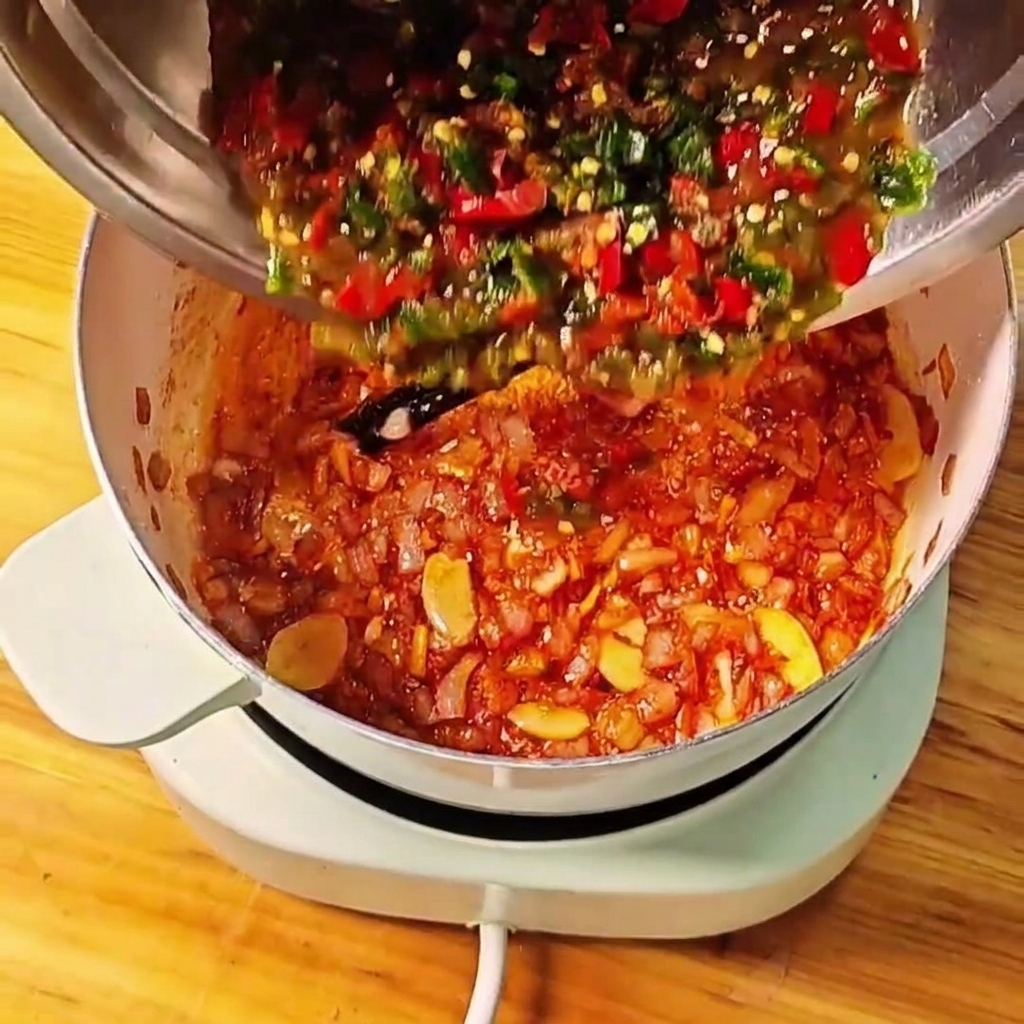 Tomates majados y chiles verdes y rojos siendo vertidos desde un cuenco de metal a la olla de cocción.