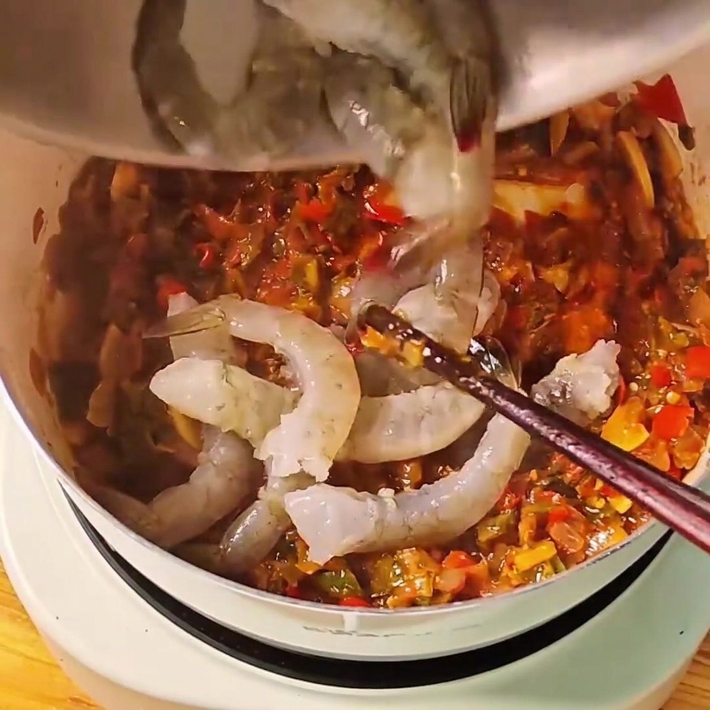 Camarones enteros grises crudos siendo arrojados a una salsa roja de tomate a fuego lento en una olla blanca.