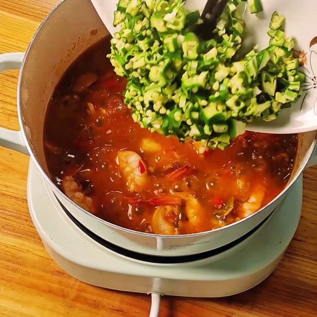 Okra verde recién picada deslizándose desde un plato blanco al estofado burbujeante.
