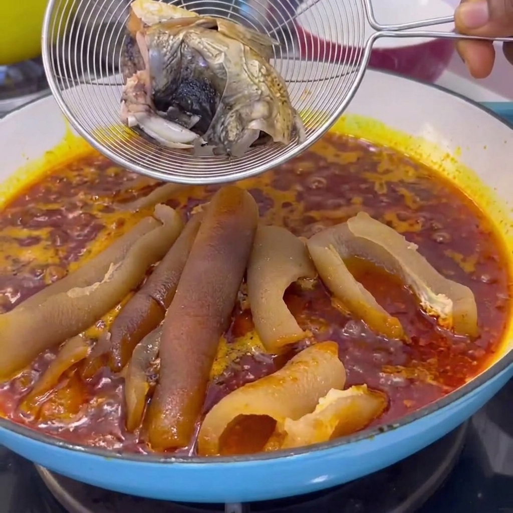 Un colador de alambre que sostiene trozos de pescado cocido siendo bajado a una olla con salsa roja hirviendo y cuero de cerdo.
