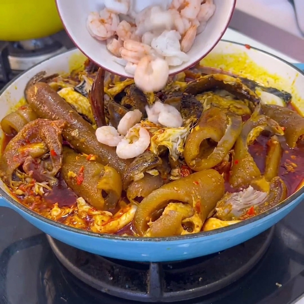 Un tazón de camarones blancos pelados siendo incorporados en una olla de cocción llena de cuero de cerdo estofado y salsa de chile.