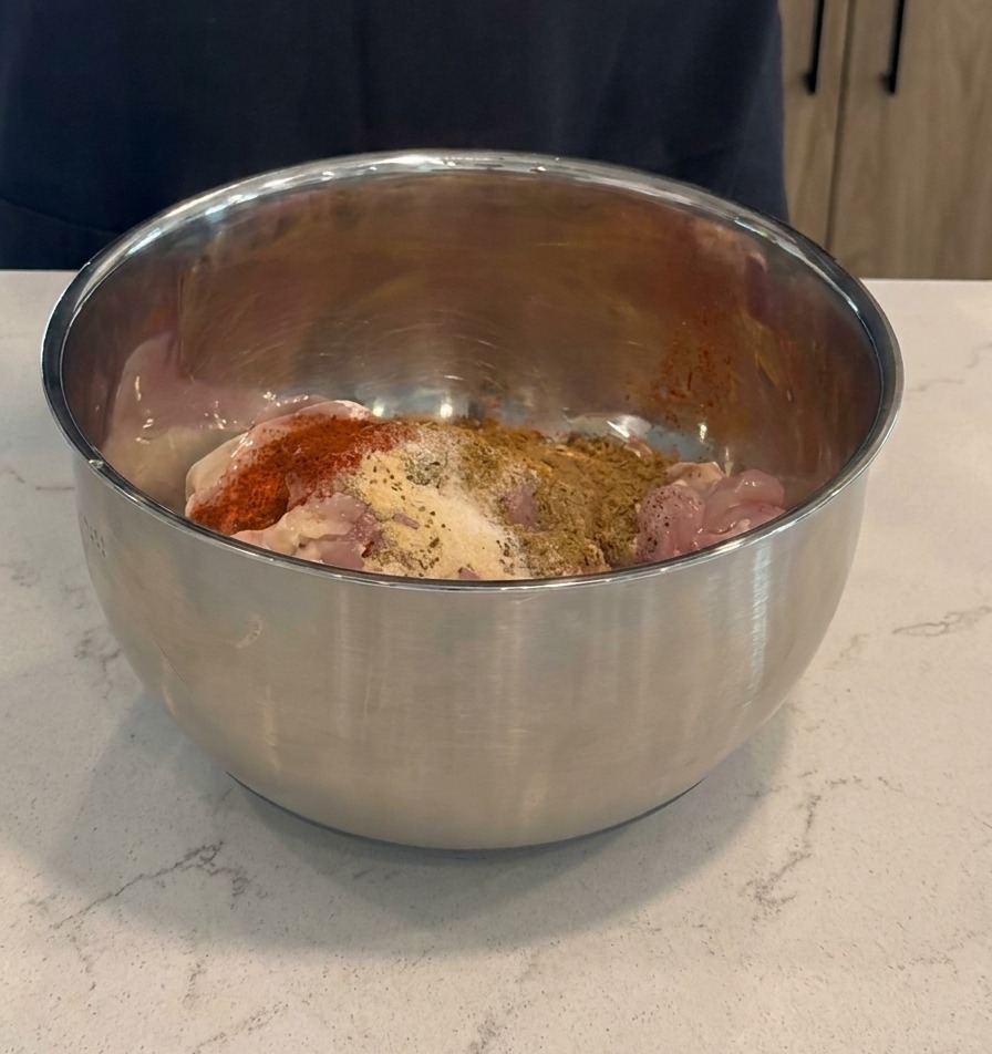 Agregando adobo rojo oscuro y especias secas a los muslos de pollo crudos dentro de un tazón de acero inoxidable.
