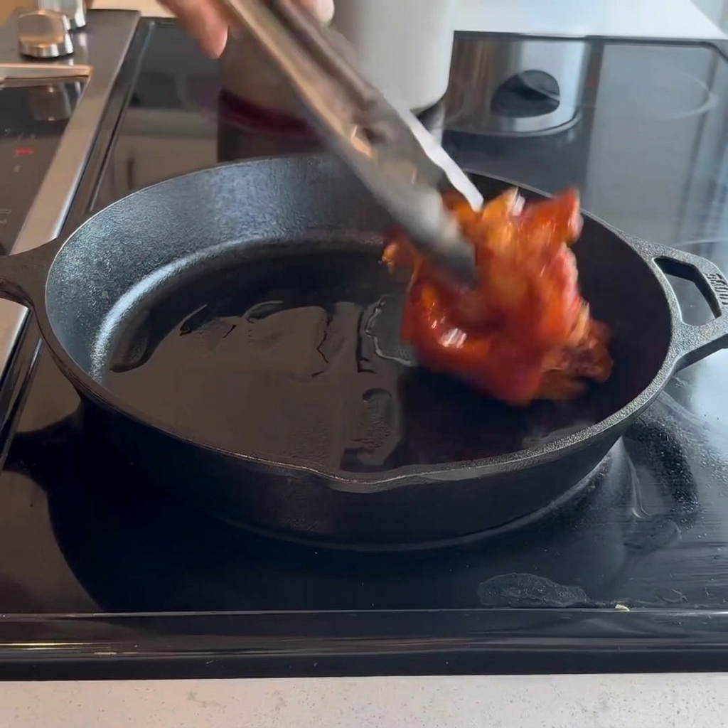 Muslos de pollo crudos marinados colocados con la piel hacia abajo en una sartén de hierro fundido caliente usando pinzas de metal.
