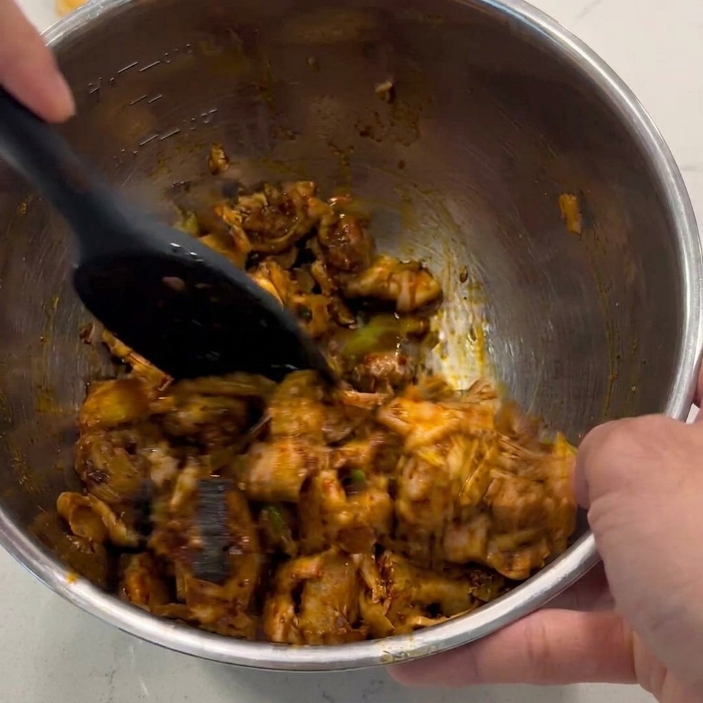Una espátula negra mezclando y revolviendo trozos de pollo para cubrirlos con una salsa marrón brillante dentro de un tazón de metal.