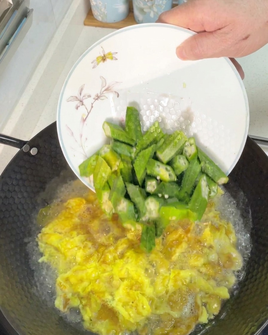 Añadiendo trozos de okra verde recién cortada en un wok con caldo de huevo hirviendo.