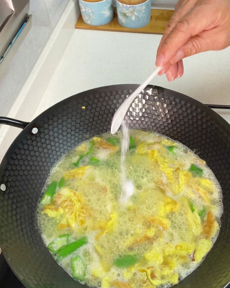 Mano añadiendo una cucharada de sal blanca a un wok de sopa de okra y huevo a fuego lento.