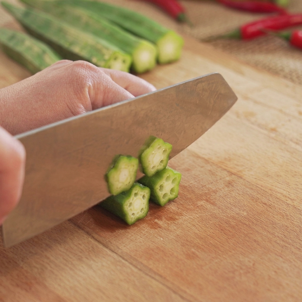 Okra verde siendo cortada en rodajas redondas en forma de estrella sobre una tabla de cortar de madera con un cuchillo de cocina.