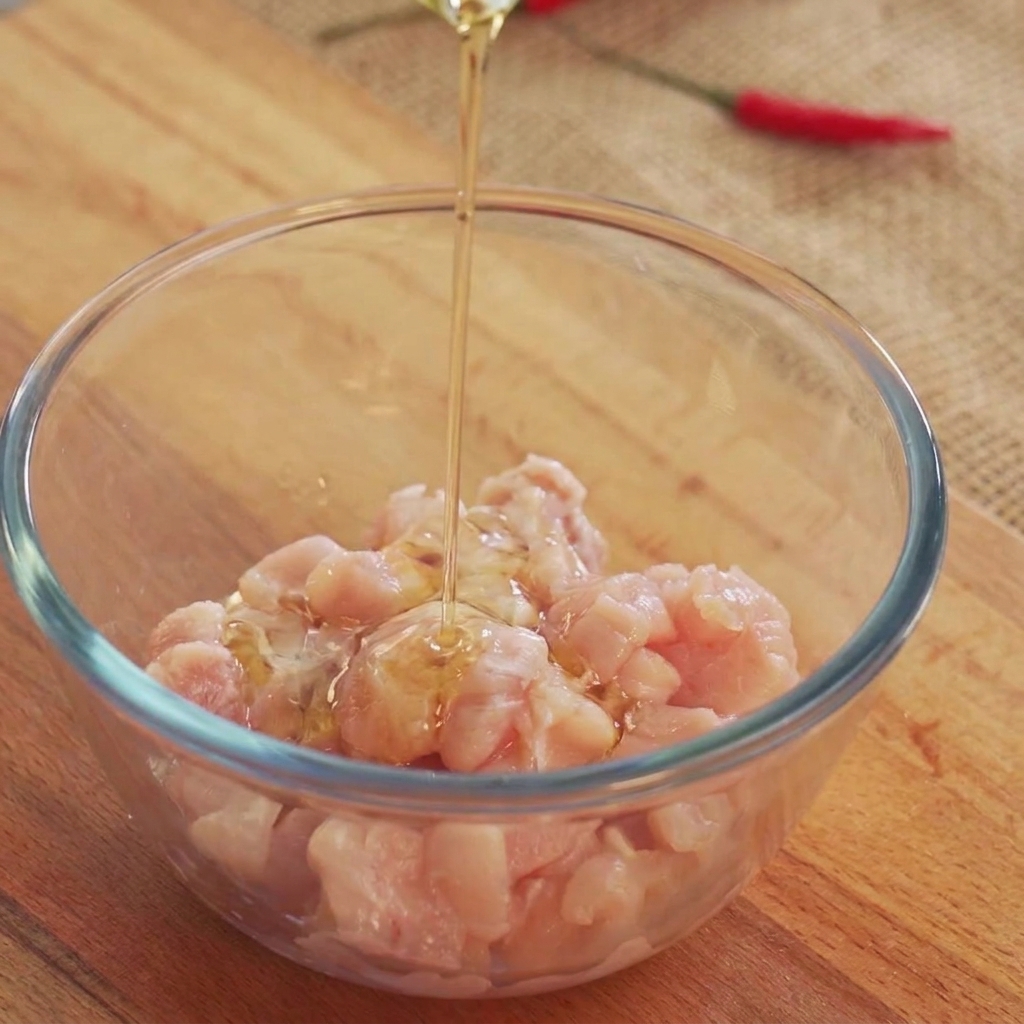 Aceite de cocina siendo vertido desde arriba sobre trozos de pollo crudo dentro de un bol de vidrio.
