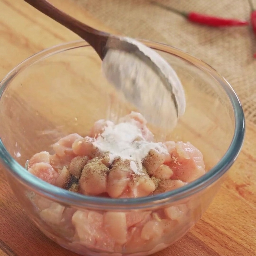 Vino de cocina y polvos sazonadores blancos siendo espolvoreados sobre cubos de pollo crudo en un bol de vidrio.