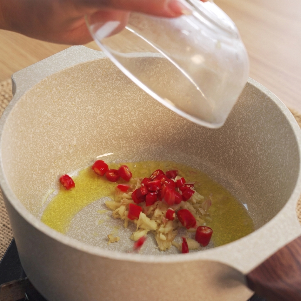 Mano dejando caer chiles rojos picados en una olla con aceite caliente y jengibre picado.