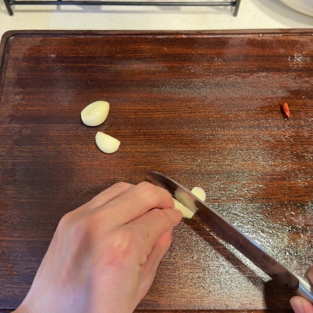 Dientes de ajo siendo cortados con un cuchillo en una tabla de madera junto a un trozo de chile rojo.