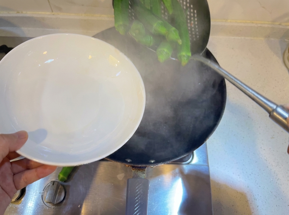 Okras verdes enteras siendo blanqueadas en una olla con agua hirviendo usando una espumadera.