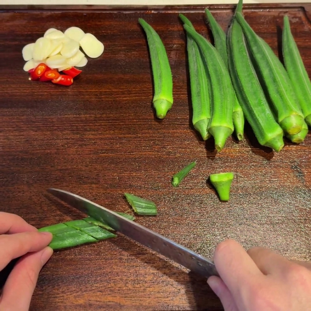 Okra verde blanqueada siendo cortada en diagonal con un cuchillo sobre una tabla de madera.
