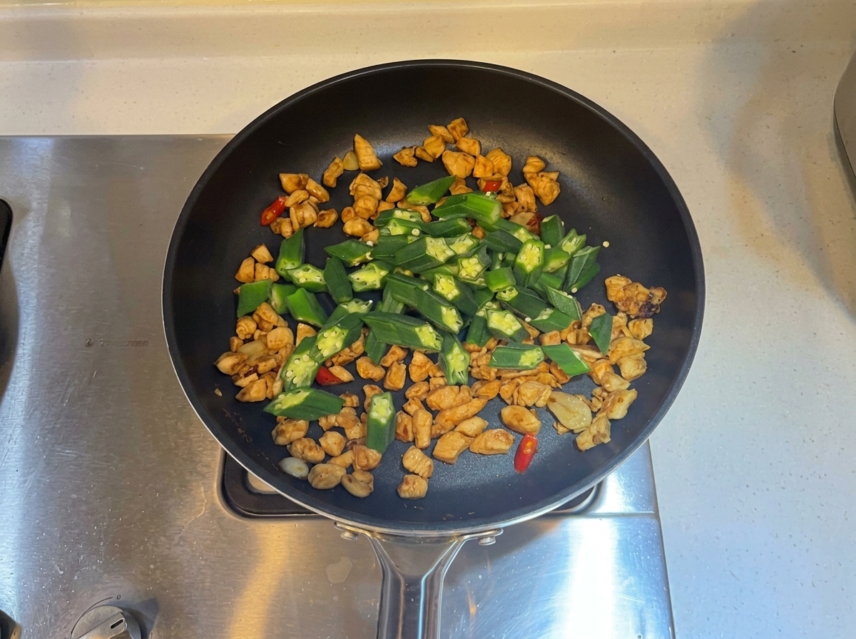 Rodajas de okra verde mezcladas en la sartén con el pollo cocinado y los aromáticos.