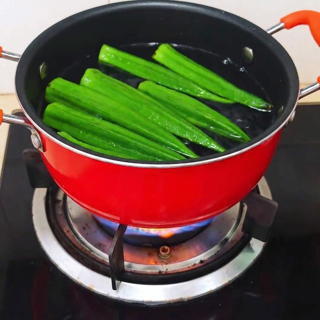Vainas de okra crudas enteras colocándose en una olla roja con agua hirviendo.