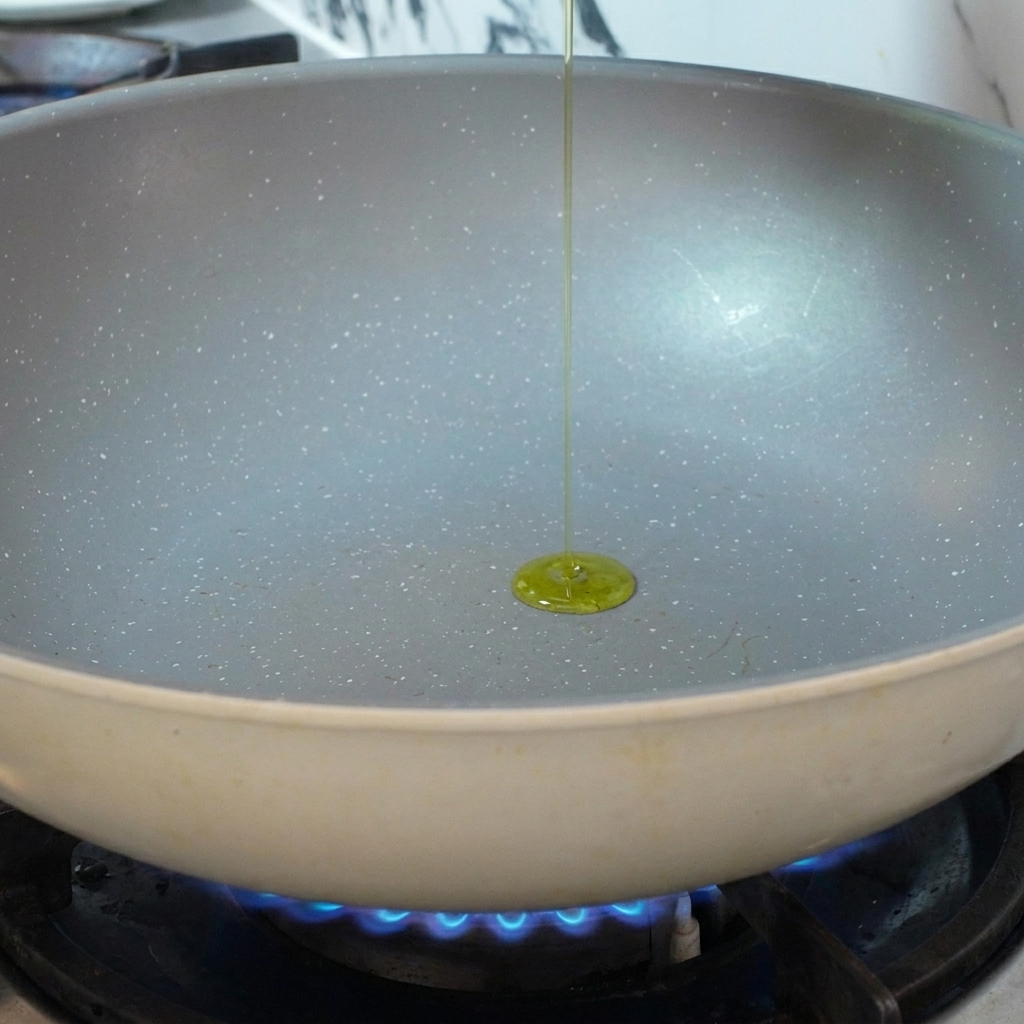 Aceite de oliva siendo vertido en una sartén de cerámica precalentada sobre una llama de gas azul.