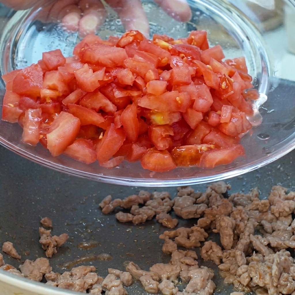 Tomates en dados y cebollas rojas siendo vertidos desde un bol de cristal a la sartén con la carne.