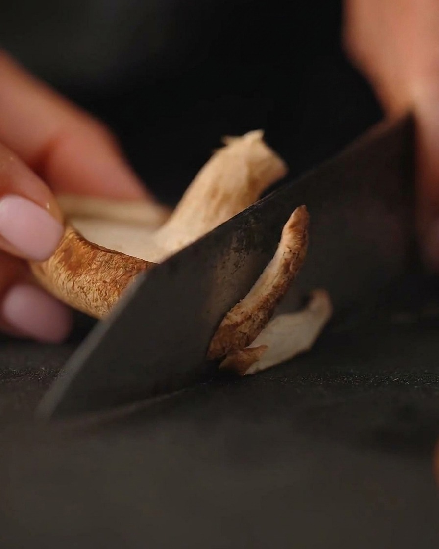 Primer plano de champiñones frescos siendo cortados en una tabla de cortar oscura con un cuchillo.