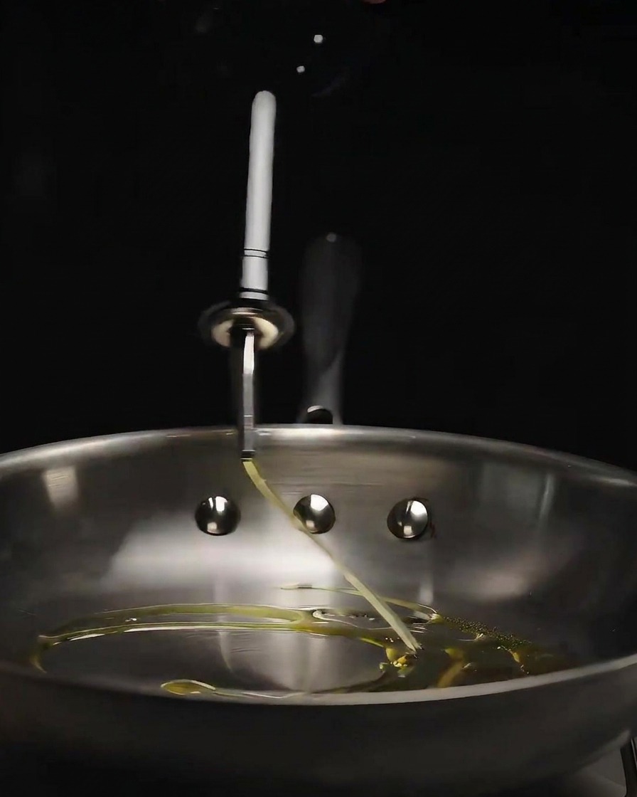 Aceite de oliva siendo vertido desde una botella de vidrio en una sartén de acero inoxidable brillante.
