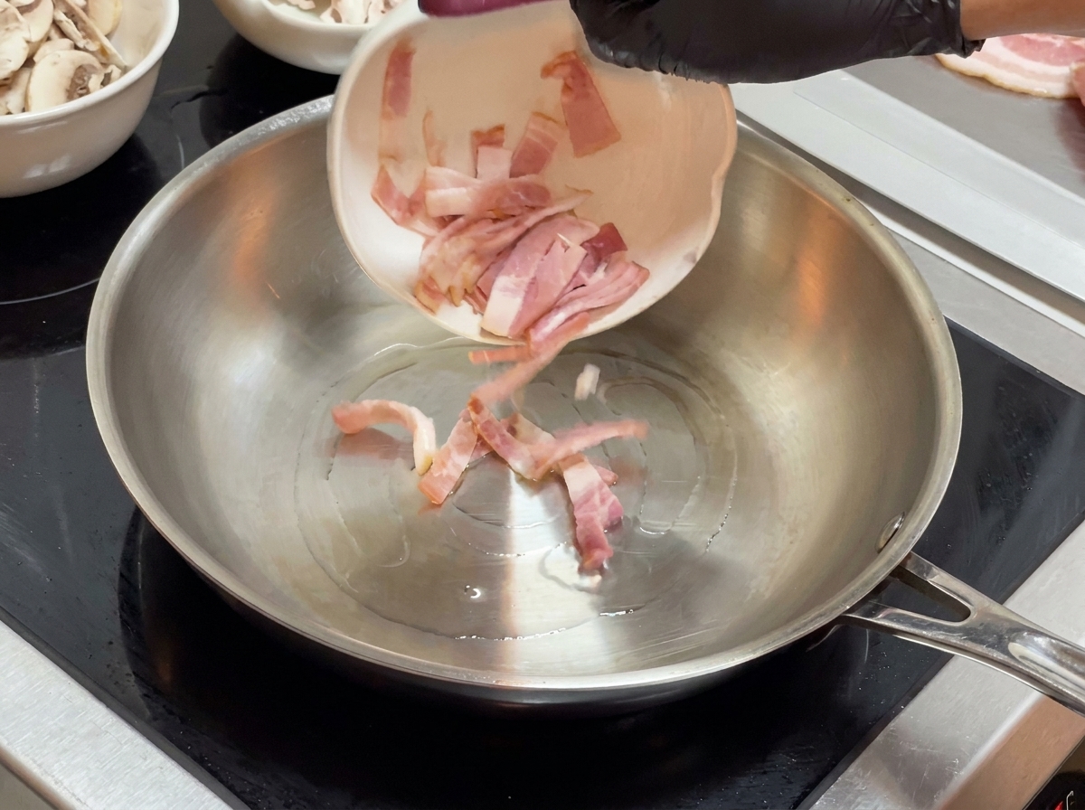 Trozos de tocino vertidos desde un pequeño bol blanco en una sartén de acero inoxidable.