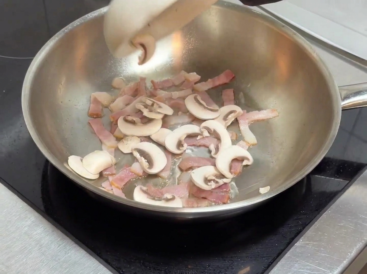 Champiñones rebanados siendo añadidos a la sartén que contiene los trozos de tocino cocinados.