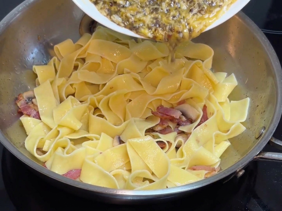 Salsa de crema de trufa negra preelaborada siendo vertida desde un plato blanco a una sartén de acero inoxidable que contiene pasta ancha.