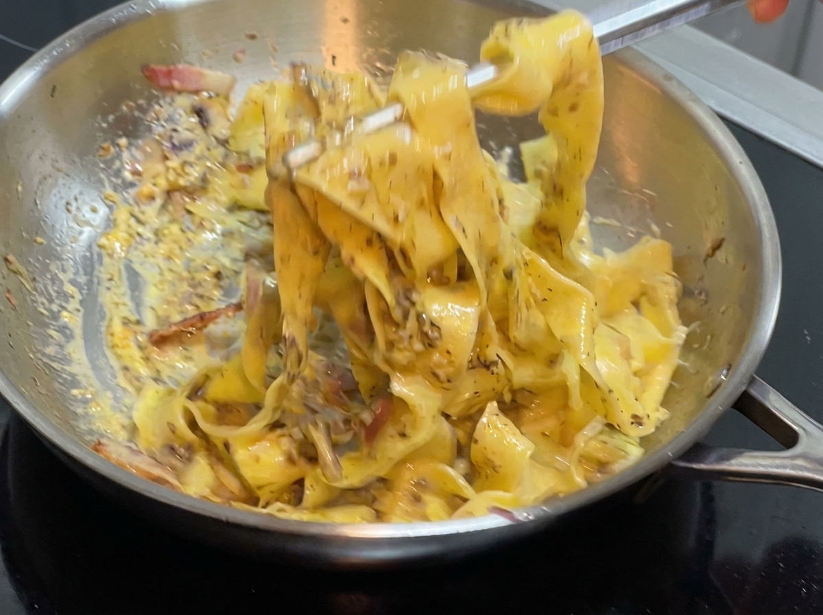 Unas pinzas mezclando pasta ancha y salsa dentro de una sartén de acero inoxidable en la estufa.