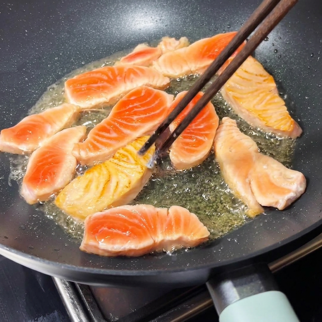 Un par de palillos de madera volteando un trozo de salmón sellado de color marrón dorado dentro de una sartén caliente.