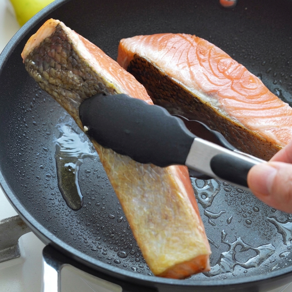 Filetes de salmón siendo volteados con pinzas negras dentro de una sartén caliente con aceite, mostrando la piel sellada de color marrón dorado.