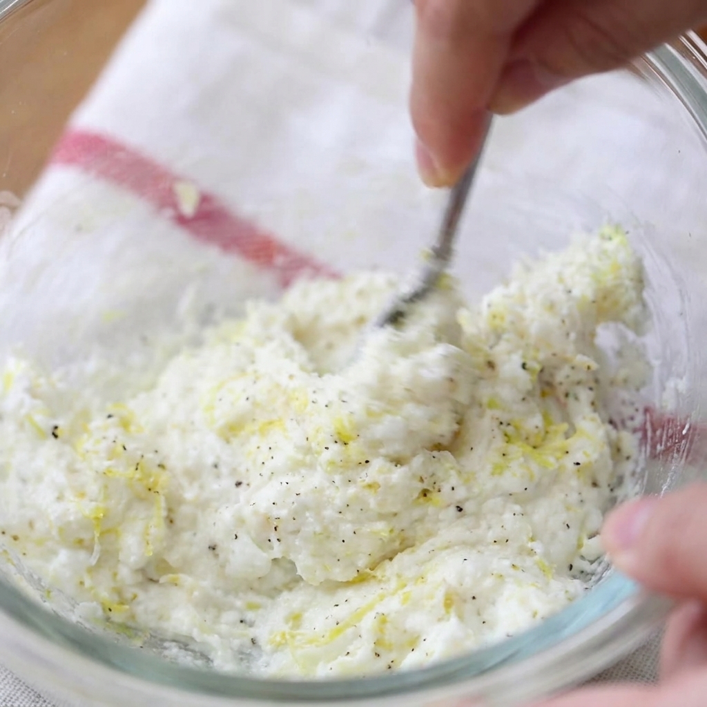 Una mano usando un tenedor para mezclar queso crema, ralladura de limón y pimienta negra en un tazón de vidrio.