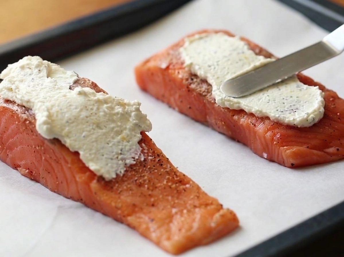 Una espátula de metal extendiendo una capa gruesa de la mezcla blanca de queso crema sobre dos filetes de salmón crudo en una bandeja para hornear forrada con papel pergamino.