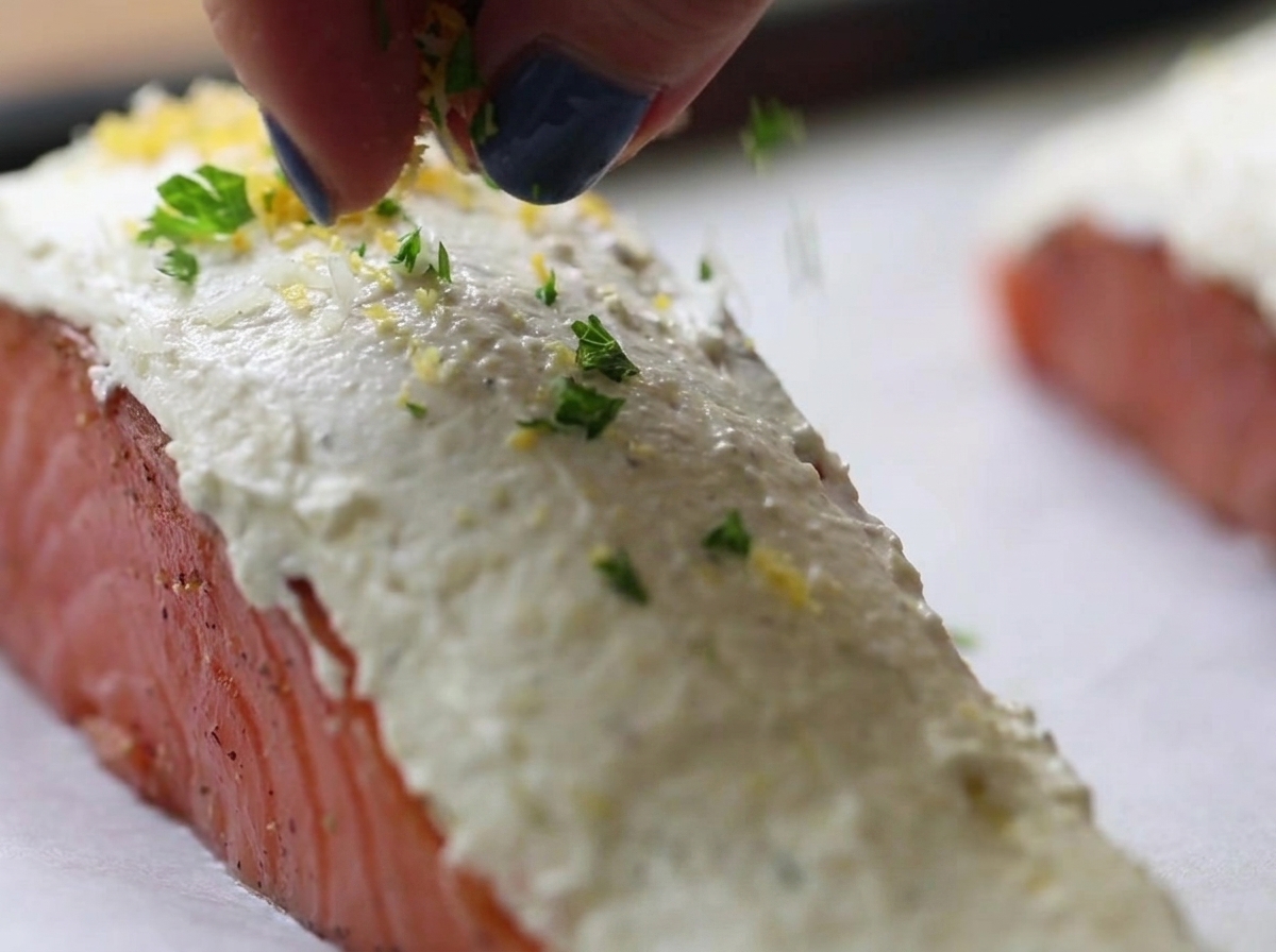 Dedos espolvoreando una mezcla verde y amarilla de hierbas y pan rallado sobre filetes de salmón crudo cubiertos de queso crema blanco.