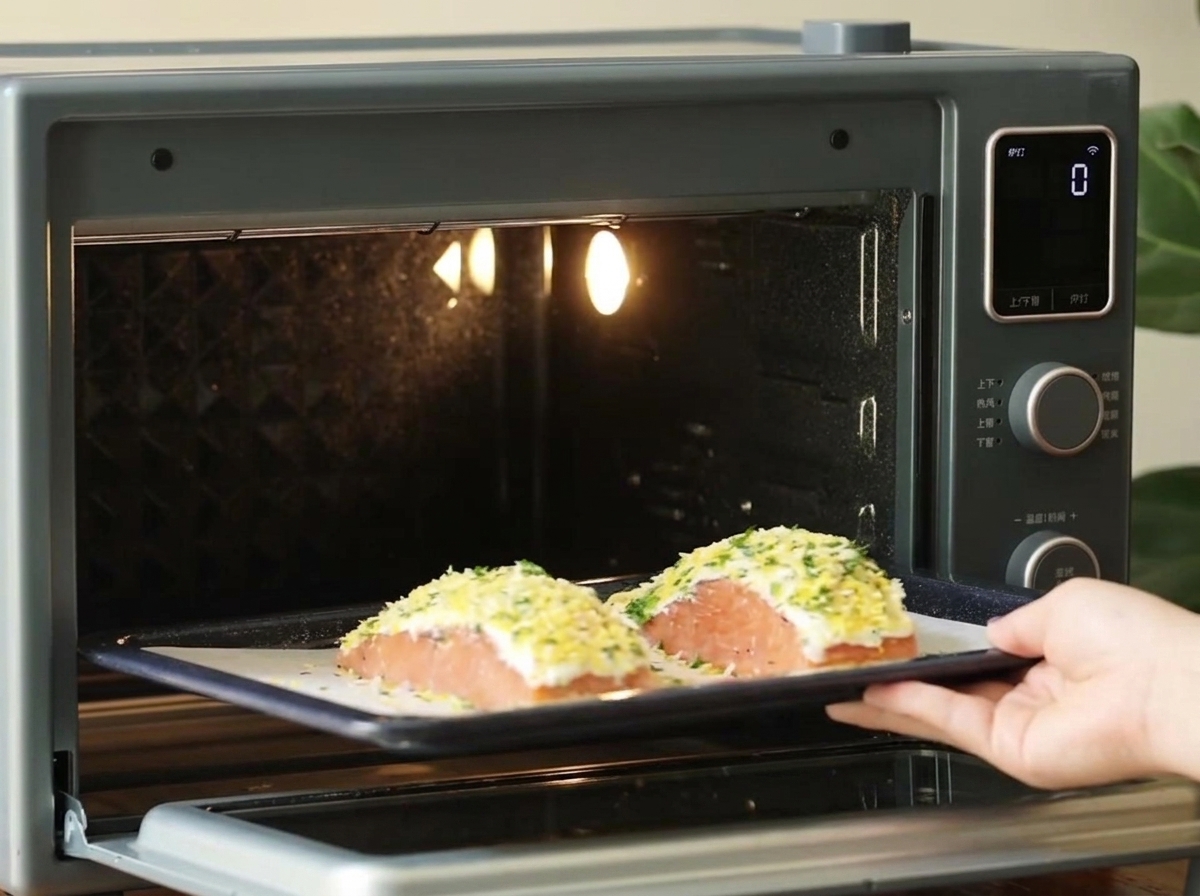 Una mano deslizando una bandeja para hornear oscura con dos filetes de salmón empanizados en un horno de mostrador pequeño y bien iluminado.